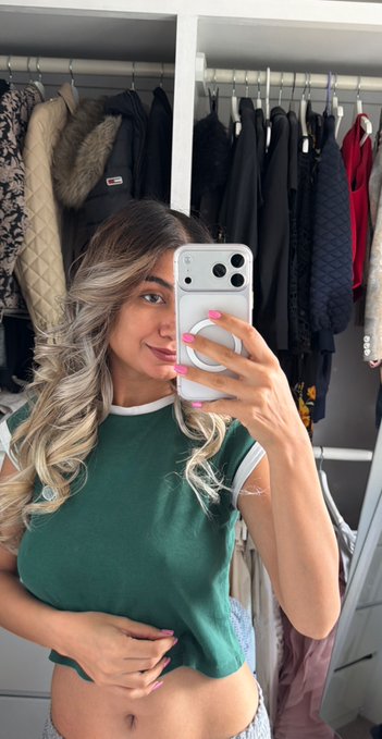 A woman with blonde hair takes a mirror selfie in her closet, wearing a green t-shirt with white trim and pink nail polish.