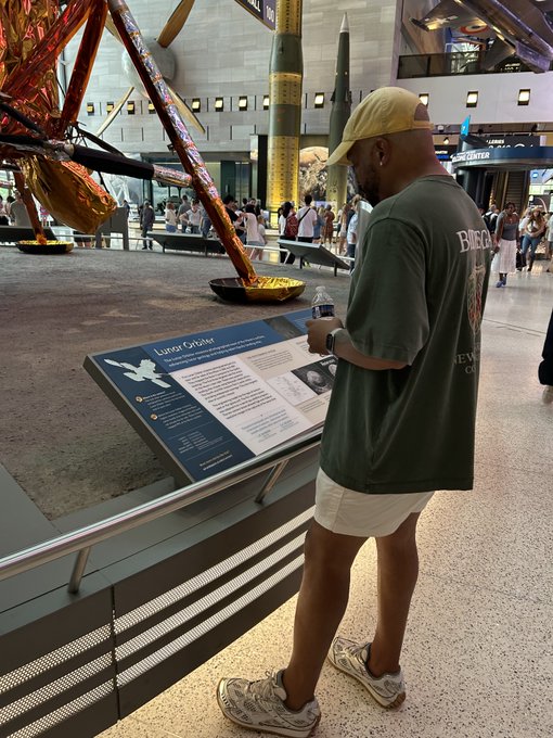 A man wearing a cap and green shirt reads an informational placard about a 'Lunar Crater' at a museum exhibit featuring rockets.