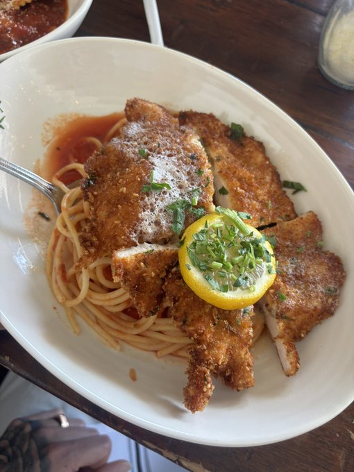 A bowl of pasta topped with breaded chicken cutlets, garnished with a lemon slice and chopped green herbs, served with marinara sauce on a wooden table.