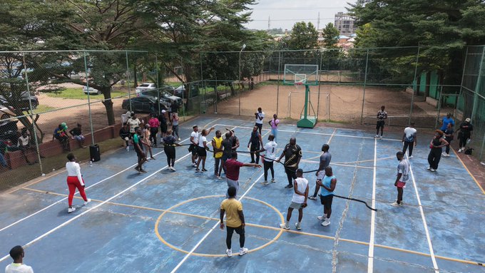 An overhead view of a group of people gathered on a blue outdoor basketball court, with a basketball hoop, chain-link fencing, and trees visible in the background.