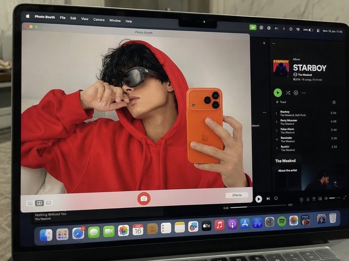 A laptop screen showing Photo Booth with a selfie of a person in a red hoodie taking a mirror photo with an orange iPhone, alongside a Spotify window playing The Weeknd's "STARBOY" album.