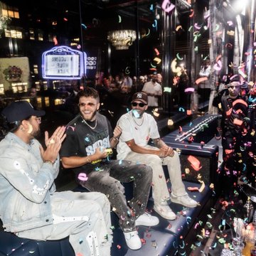 Three men celebrating amid falling confetti in a dimly lit venue, possibly a casino or upscale lounge, with one wearing sunglasses and a chain.