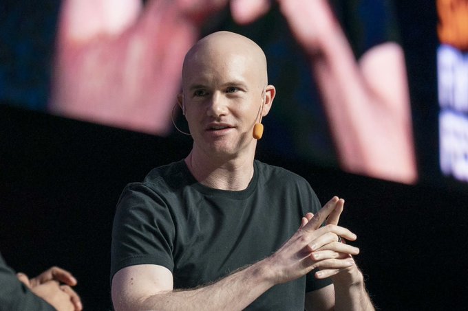 A bald man in a black t-shirt speaks on stage with a headset microphone, his hands clasped together in front of a dark background with blurred pink shapes.