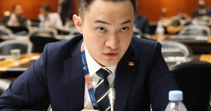 A young Asian man in a navy suit and striped tie sits at a formal conference table with a microphone, wearing a lanyard and a "TRON" lapel pin, appearing focused during a panel discussion. The caption suggests he is presenting TRON's blockchain achievements, specifically highlighting its 3.93 million daily active users as evidence of the network's global dominance.