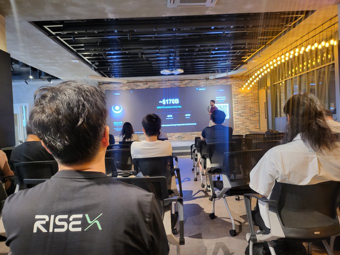View from behind an audience attending a presentation in a modern venue; one attendee wears a shirt with "RISEX" on the back, while a projection screen shows "~$170B".