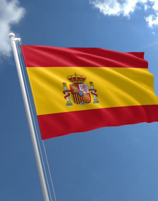 The image shows the flag of Spain waving against a blue sky, featuring its iconic red and yellow horizontal bands with the Spanish coat of arms in the center. The flag visually represents the country's latest regulatory development, as the accompanying news announces Spain's confirmation that self-custody crypto holdings are exempt from foreign asset disclosure requirements.