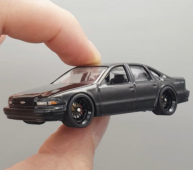 A miniature black Chevrolet Impala model car being held between a person's fingers, showcasing detailed wheels and a lowered stance.