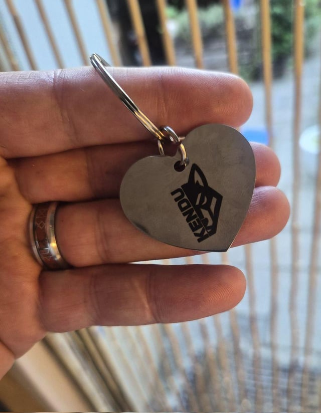 A hand displays a metal heart-shaped keychain engraved with the "KENDU" logo, showcasing a physical example of the project's branding. The keychain ties directly into the post's message about making "$KENDU visible everywhere."