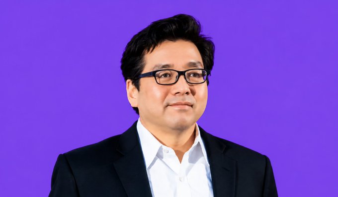 Professional headshot of an Asian man wearing glasses and a dark suit jacket over a white shirt against a purple background.