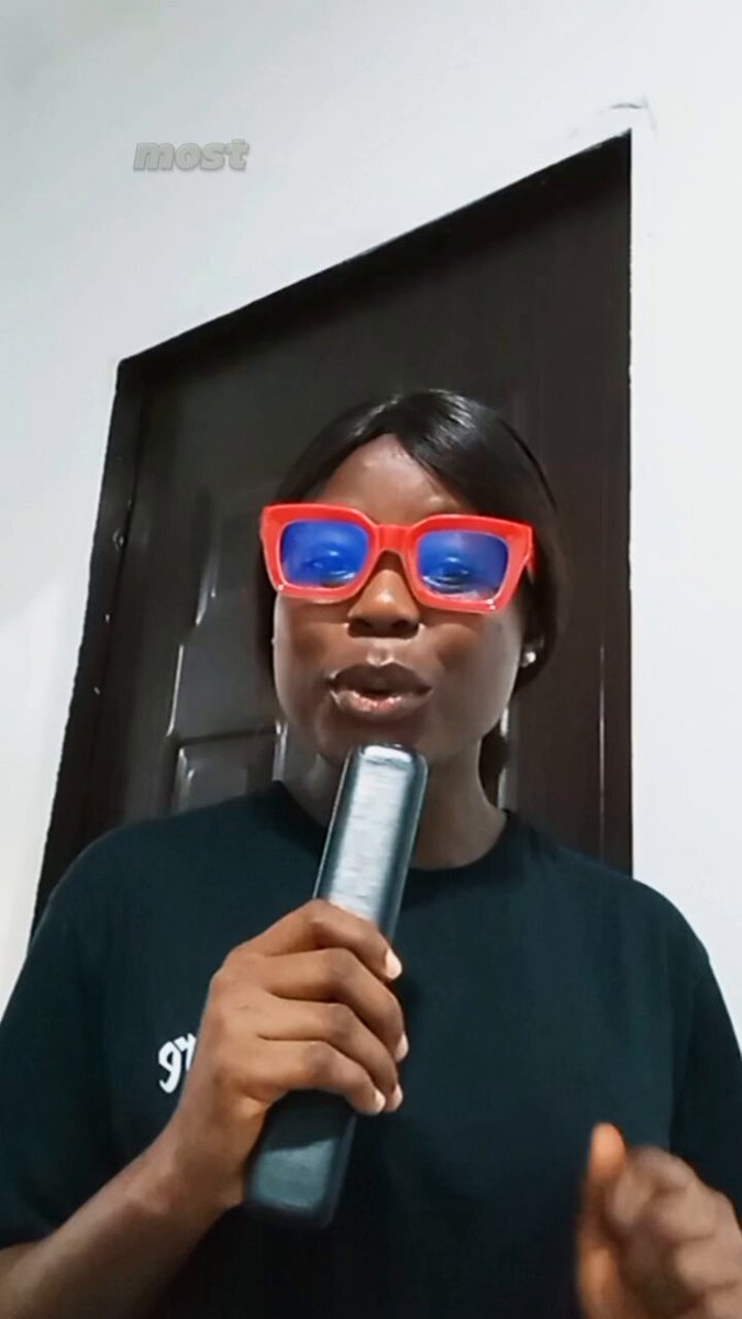 The image features a person wearing red glasses and holding a remote control. The person appears to be talking into a microphone or speaking into a camera, possibly giving a speech or presentation. The background of the image shows a door with a window behind it, suggesting that they are in an indoor setting like a room or office.