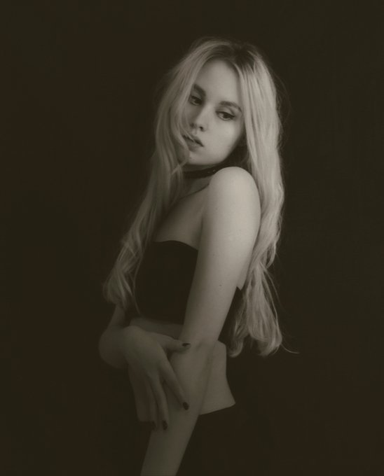 A black and white portrait of a young blonde woman wearing a black strapless top and choker, posing against a dark background with long hair cascading down.