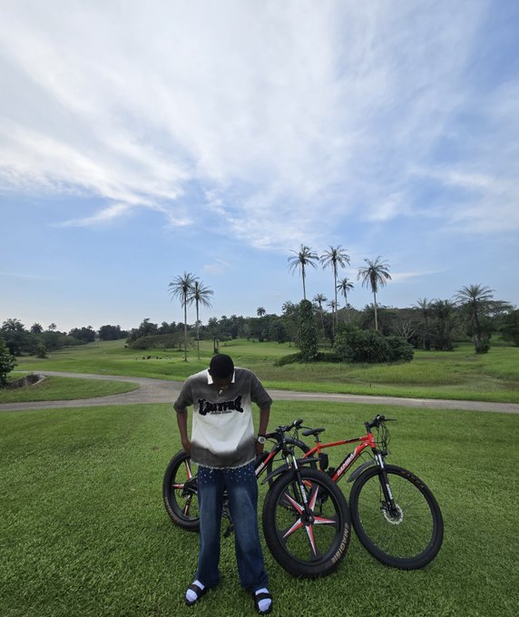 A person wearing a Lakers jersey and face mask stands next to a red and black mountain bike on a grassy field with palm trees in the background.