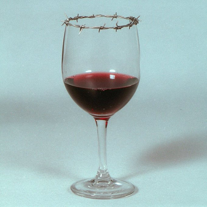A conceptual photograph showing a wine glass filled halfway with red wine, with a loop of barbed wire wrapped around the rim of the glass against a light blue background.