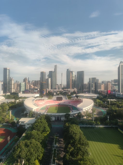 An aerial view of a city skyline featuring modern skyscrapers behind a large oval sports stadium with red and white seating, surrounded by green trees under a partly cloudy blue sky.