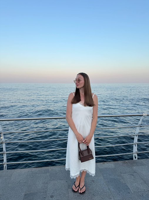 A young woman in a white sundress and sunglasses stands on a seaside promenade holding a small brown handbag, with the ocean and sky behind her.