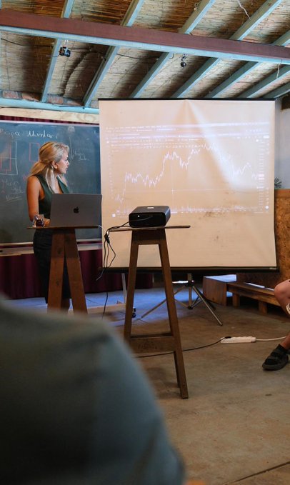 A blonde woman in a black dress stands beside a laptop and projector, presenting a chart with fluctuating line graphs on a large screen in a rustic classroom. The setting, with chalkboards and wooden beams, aligns with the post about engaging students at Rlay Blockchain Week 2026 in Şirince.