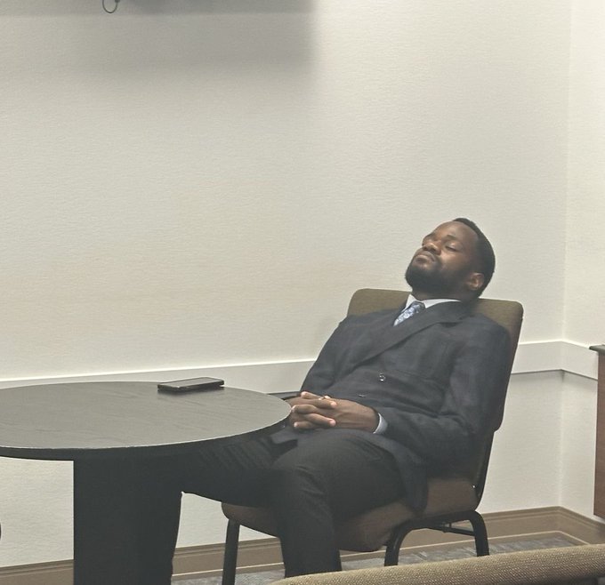A man in a dark double-breasted suit with a blue tie appears to be sleeping or resting with his eyes closed while seated at a round table with a phone placed in front of him.