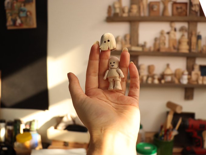 A hand holds up two tiny wooden figurines—a smiling doll and a small character with a cape of hair—in what appears to be a woodcarver's workshop, with shelves full of similar handcrafted pieces visible in the background. The scene ties to the "Gm friends" greeting by showcasing the friendly, morning charm of handmade artistry.