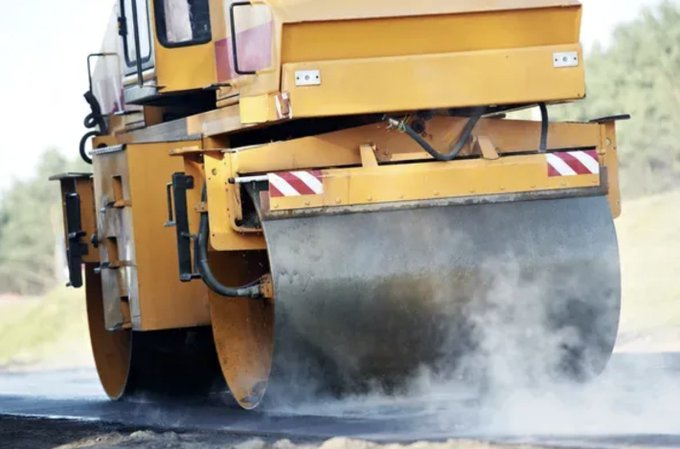 A yellow steamroller compacts a dark asphalt road, emitting white steam or dust from the surface.