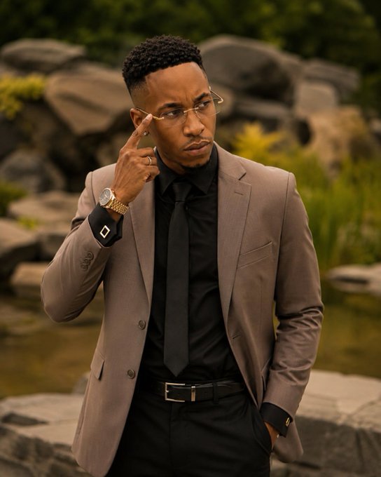 A young man wearing a tan suit, black shirt, and tie, posing outdoors with gold-rimmed glasses and a gold watch.