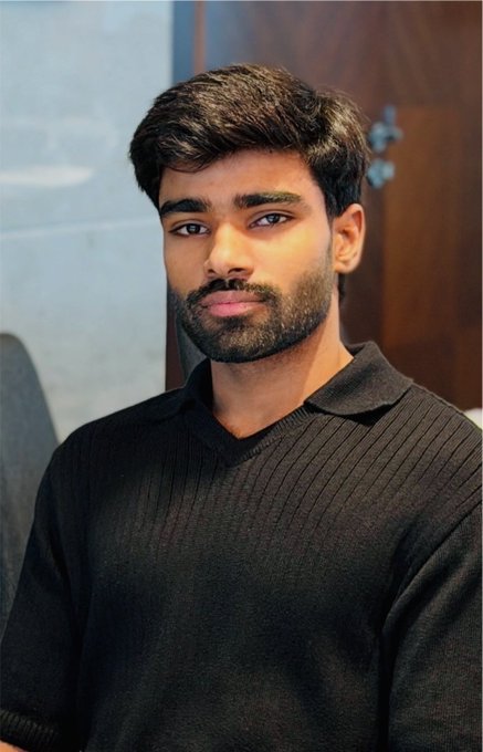 A portrait photo of a young man with dark hair and a short beard, wearing a black polo-style sweater, posed indoors.