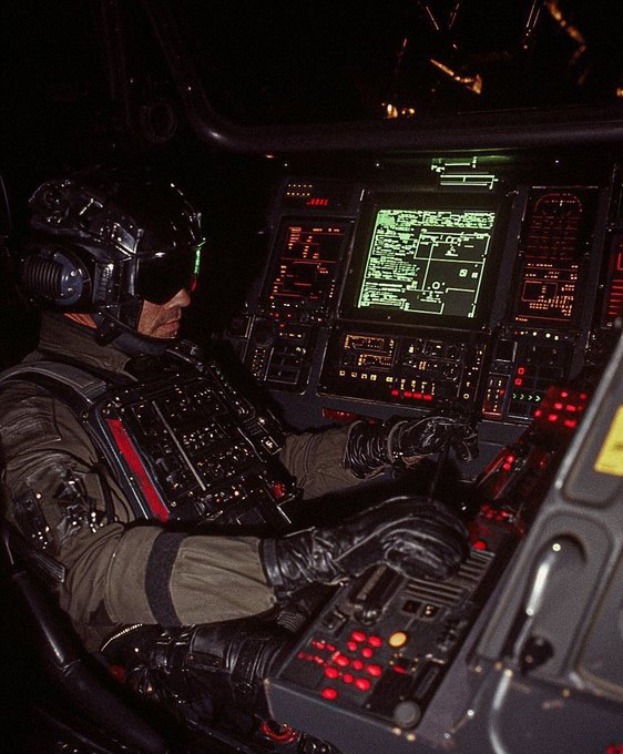 A pilot in a high-tech helmet and flight suit sits at the controls of a futuristic aircraft cockpit filled with illuminated displays and buttons.