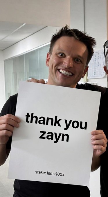 A man smiles excitedly while holding a white sign that reads 'thank you zayn' with 'stake: lemz100x' printed at the bottom.