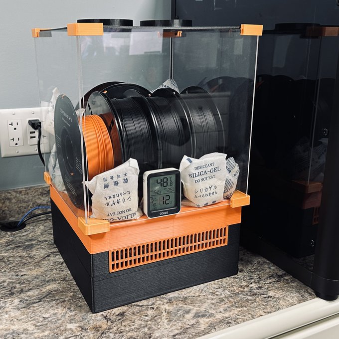 A filament dryer box containing orange and black 3D printing filament spools, with a digital hygrometer displaying 48% humidity and 75°F/29°C temperature, alongside silica-gel desiccant packets, humorously repurposed as a climate-controlled Bitcoin mining rig setup.