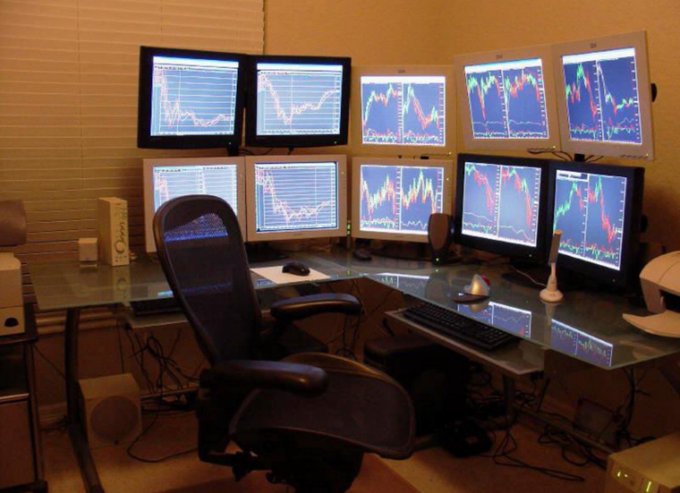 A multi-monitor trading workstation featuring eight displays arranged in two rows showing financial charts, paired with an ergonomic office chair at a glass desk.
