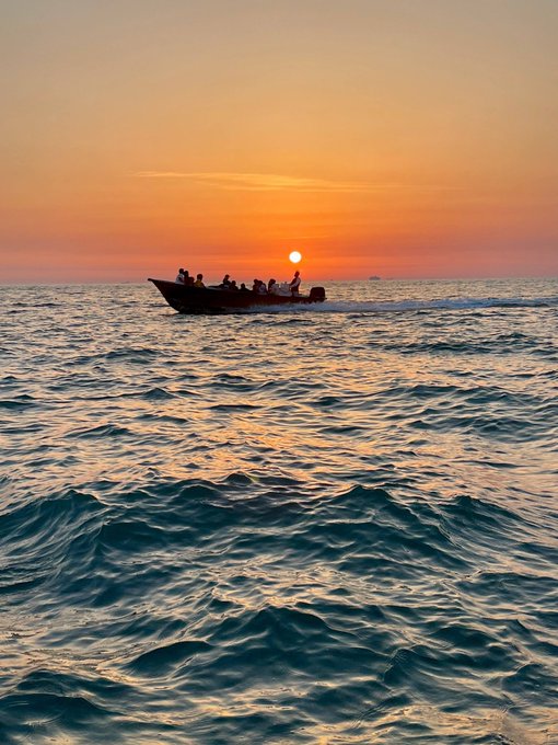 A silhouette of a boat with several passengers is visible on the ocean against a vibrant orange and yellow sunset, with the sun low on the horizon.