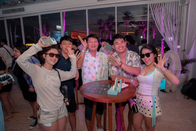 A group of friends pose playfully at a high-energy afterparty, flashing peace signs and waving by a round table with event menus, set against a backdrop of palm trees and pink-lit windows at what appears to be a rooftop venue. The celebratory scene ties directly to the "UEX Global Alpha Tournament" afterparty, capturing the victorious and social spirit of the event's closing moments.