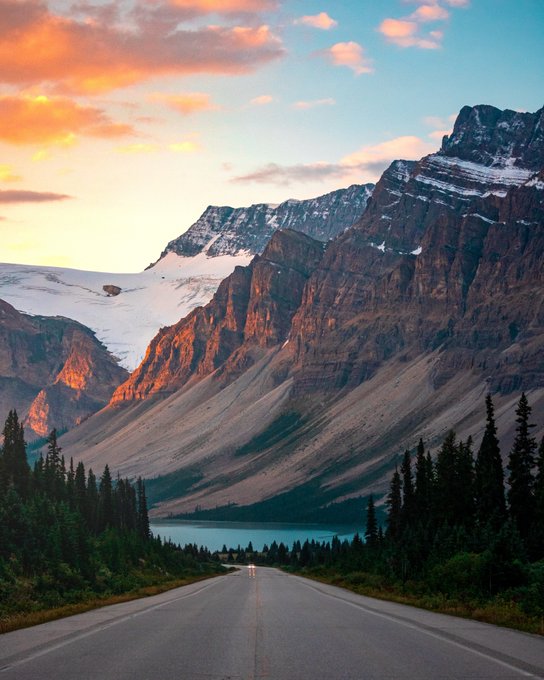 The image shows a road that leads up to a mountain range with snow-capped peaks at sunset. The sky above the mountains has a beautiful orange hue, creating a stunning backdrop for the scene. In the foreground of the photo, there is a white road stretching out into the distance, leading towards the majestic mountains.