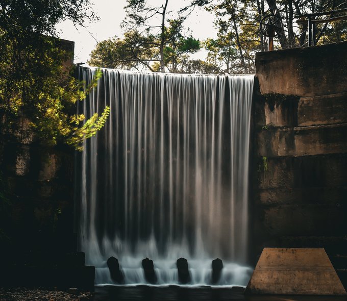 A long-exposure photograph of water cascading down a concrete dam into a pool below, surrounded by trees and stone structures.