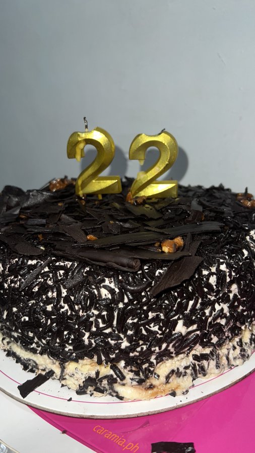 A close-up of a chocolate cake topped with chocolate shavings and two gold number '2' candles, likely for a 22nd birthday, from 'caramia.ph'.