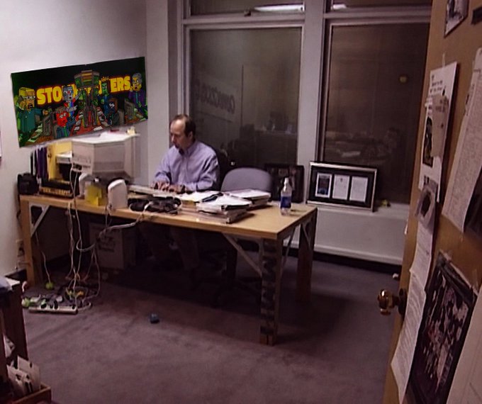 A man works at a desk in a small office with a CRT monitor and a painting on the wall featuring the text 'STORYTELLERS'.
