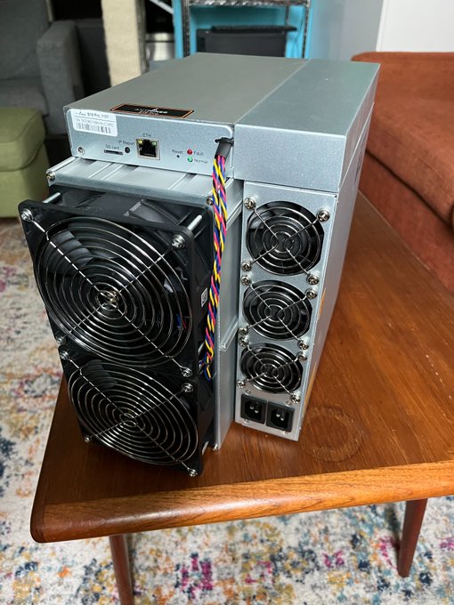 Two cryptocurrency mining machines (ASIC miners) placed on a wooden coffee table, showing their cooling fans and power supply units.
