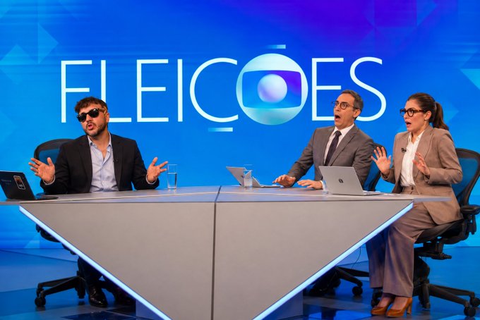 Three people sit at a sleek white desk on the set of the TV news program "FILEIÇÕES," each wearing sunglasses and displaying exaggerated, humorous expressions of surprise, with their mouths open in shock or mid-sentence. The man on the left gestures animatedly with both hands while speaking, mirroring the dramatic style suggested by the "SBT-style grilling" reference, as the woman on the right also reacts with wide-eyed astonishment, all appearing to be in the middle of a lively, theatrical on-air debate.