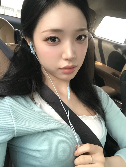 A woman with long black hair takes a selfie in a car wearing earphones, a light blue cardigan, and a seatbelt.