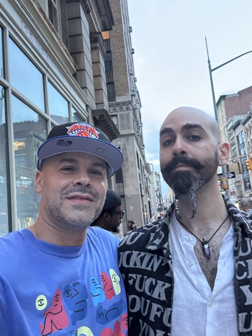 Two men pose for a selfie on a city street; one wears a New York Knicks cap and a colorful shirt, while the other sports a beard, a black and white patterned shirt, and a pendant necklace.