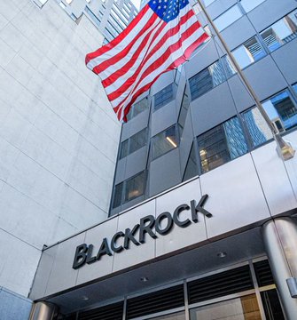 The image shows a large building with an American flag flying above its entrance. The text "BLACKROCK" is displayed prominently at the top of the building's facade, indicating that this is the name or headquarters of BlackRock, a financial services company. The presence of the American flag suggests that the company may have ties to the United States or operates in an environment where the flag holds significance.
