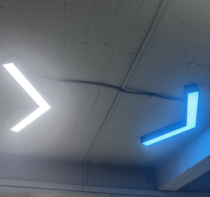 Two angular LED light fixtures mounted on a concrete ceiling, one glowing white and the other glowing bright blue.