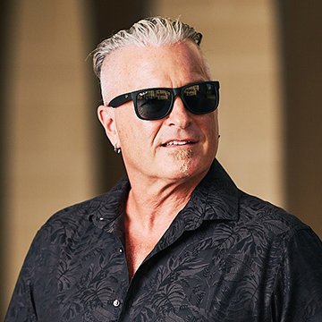 A man with short gray hair, wearing dark sunglasses and a black patterned button-up shirt, poses for a portrait photo. The accompanying post humorously suggests the man pictured is Calvin Ayre, the founder of Bodog, known for resisting Apple's newer smartphone releases in favor of his iPhone 13.