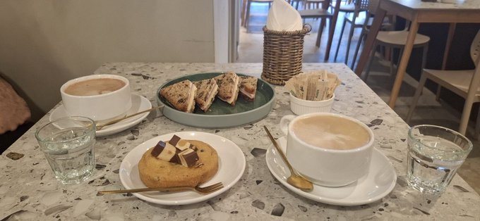 A terrazzo café table set with two cappuccinos, water glasses, a pastry topped with cream and fruit, and a plate of sliced sandwiches.