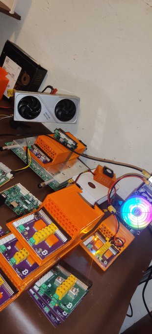 A collection of orange 3D-printed electronic housings and circuit boards (including what appears to be cryptocurrency miners) are arranged on a wooden surface next to a GPU.