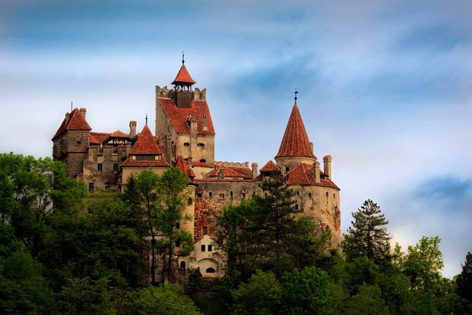 Bran Castle, a medieval fortress with distinctive red-roofed towers, stands prominently atop a forested hill against a dramatic cloudy sky.