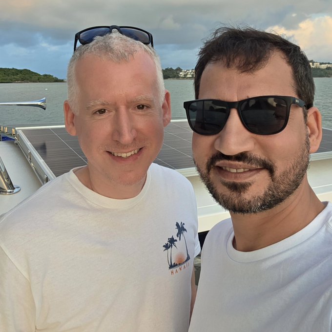 A selfie of two smiling men wearing sunglasses on a boat, with one wearing a white 'HAWAII' t-shirt, set against a scenic waterfront with buildings and a cloudy sky.