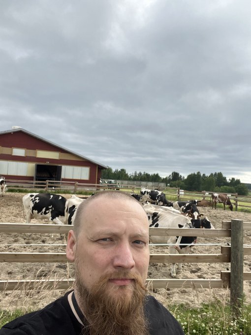 A bearded man takes a selfie in front of a farm with a red barn, where several black and white Holstein cows are gathered in a fenced enclosure under a cloudy sky.