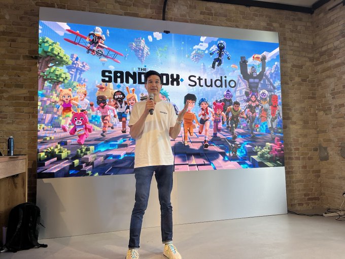 The image features a man standing in front of a large screen displaying a video game. He appears to be giving a presentation or speech about the game. The man is wearing a white shirt, blue jeans, and yellow shoes. The background shows a brick wall with various images related to the game displayed on it.