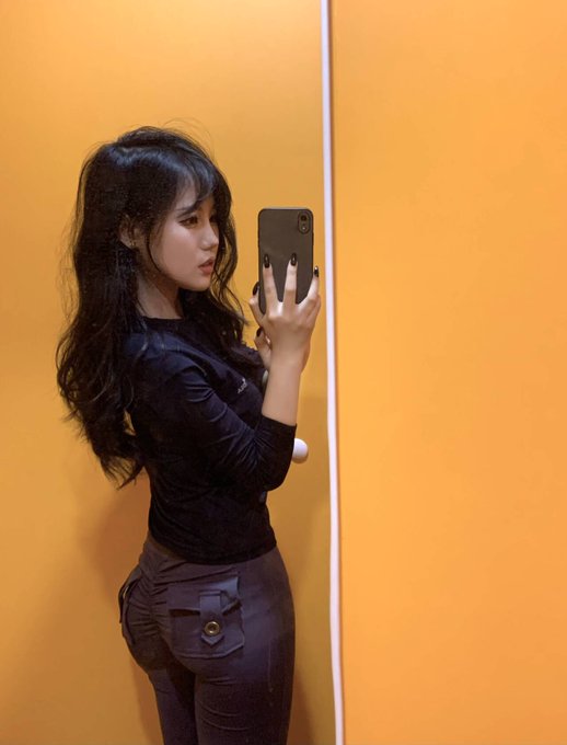 A young woman with long dark hair takes a mirror selfie in an orange-walled space, wearing a fitted black long-sleeve top and dark pants. The caption humorously compares the perkiness of her butt to the price stability needed to support Bitcoin.