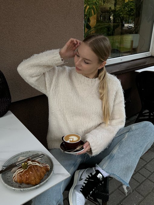 A young woman with blonde hair sits outdoors at a cafe table, wearing a fluffy white sweater and holding a cup of coffee. A pastry sits on a plate on the table in front of her.